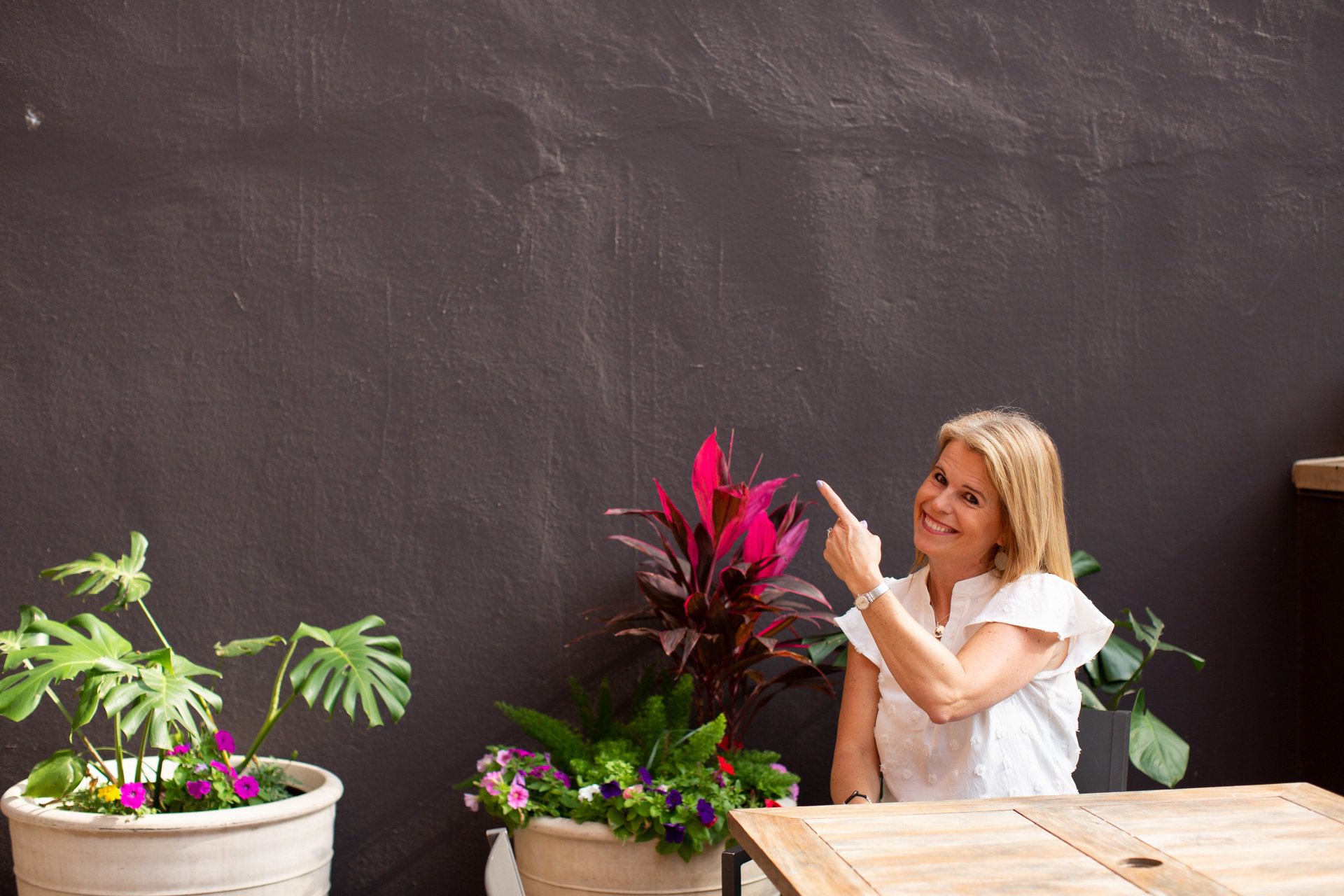Laura nelson sitting a a wood table, in front of a dark wall and a pink flower, pointing upwards and smiling