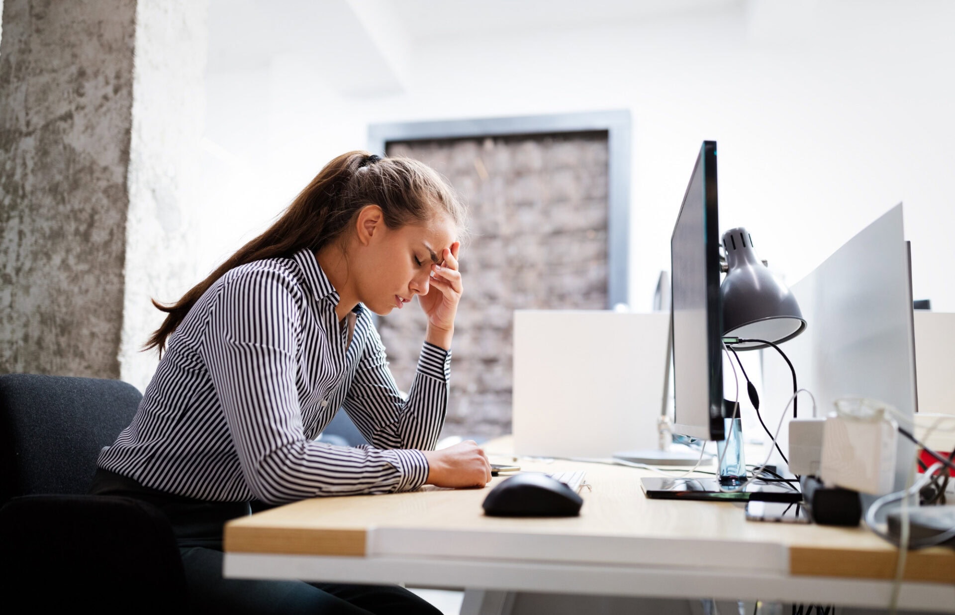 stressed out front desk worker with her head in one hand, looking down at her desk instead of at her monitor