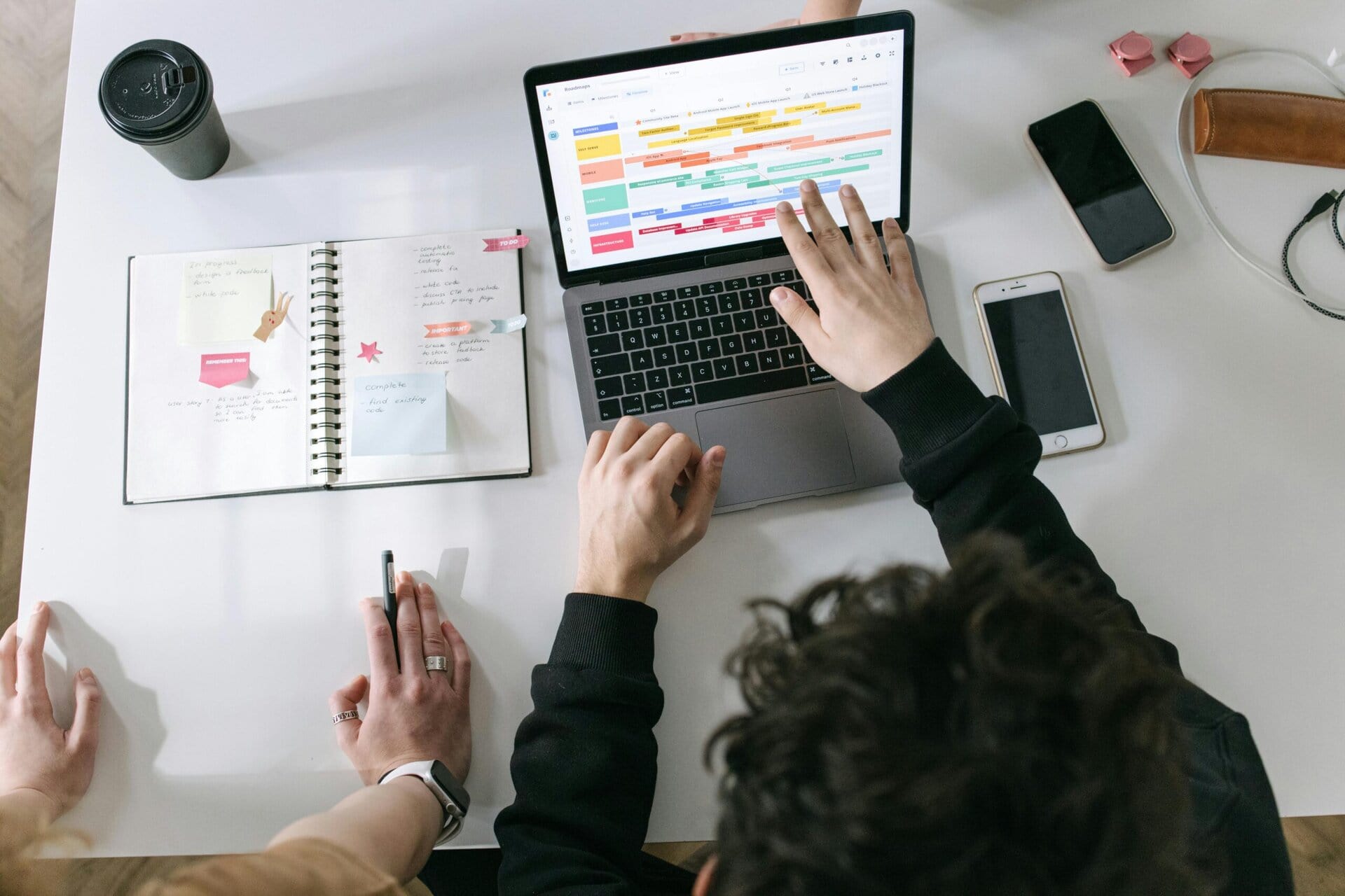 Two people looking at a laptop and refining a busy schedule. There is a notebook to the right with notes and stickers. Provided by pexels.