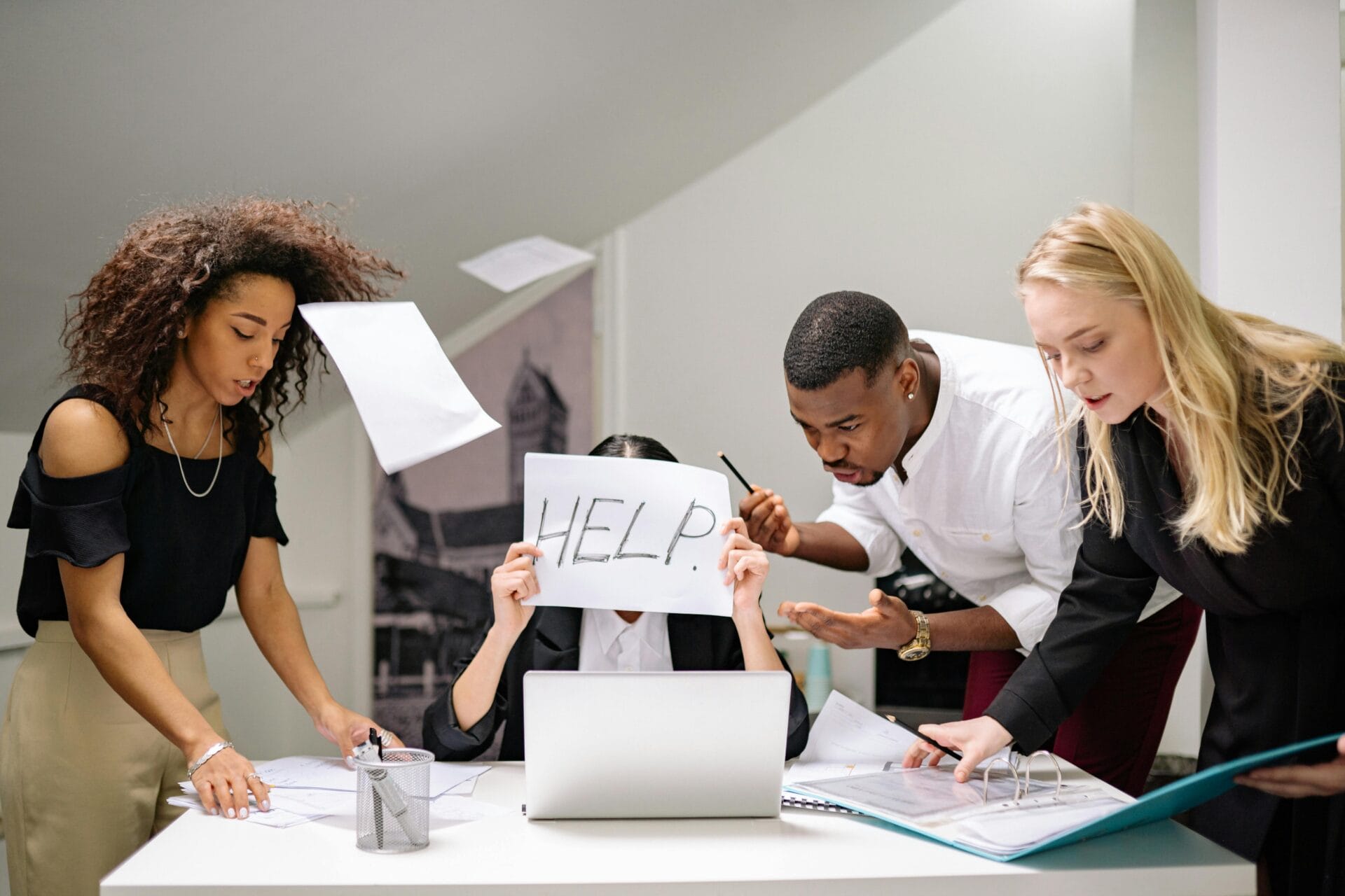 People crowded around a laptop looking stressed while one person holds a sign that says “help,” representing schedule overload and disorganized workflows.