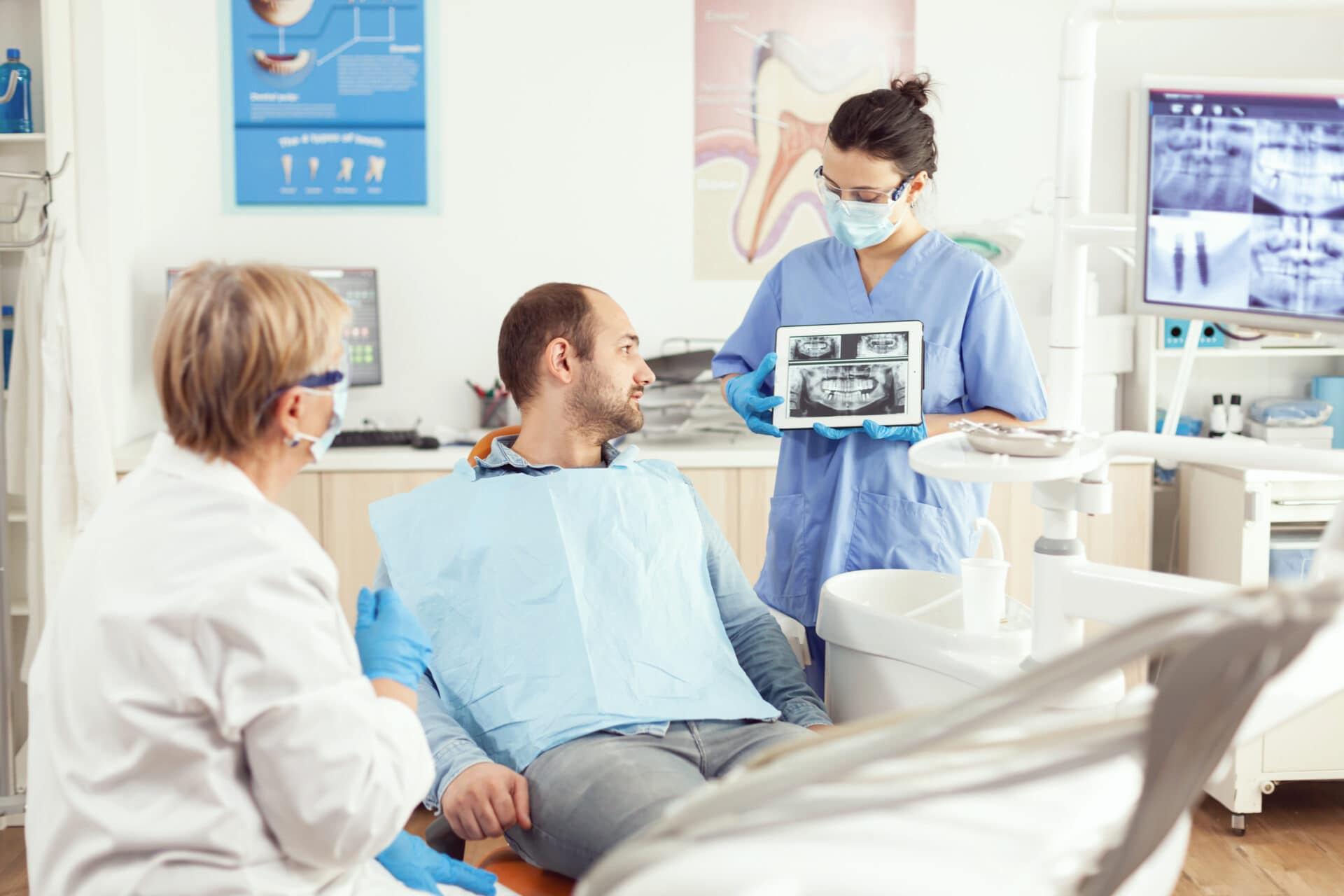 Sick man sitting on stomatological chair listening doctor while looking at tablet in dental clinic. Dentistry doctor showing to patient teeth radiography during dental consultation